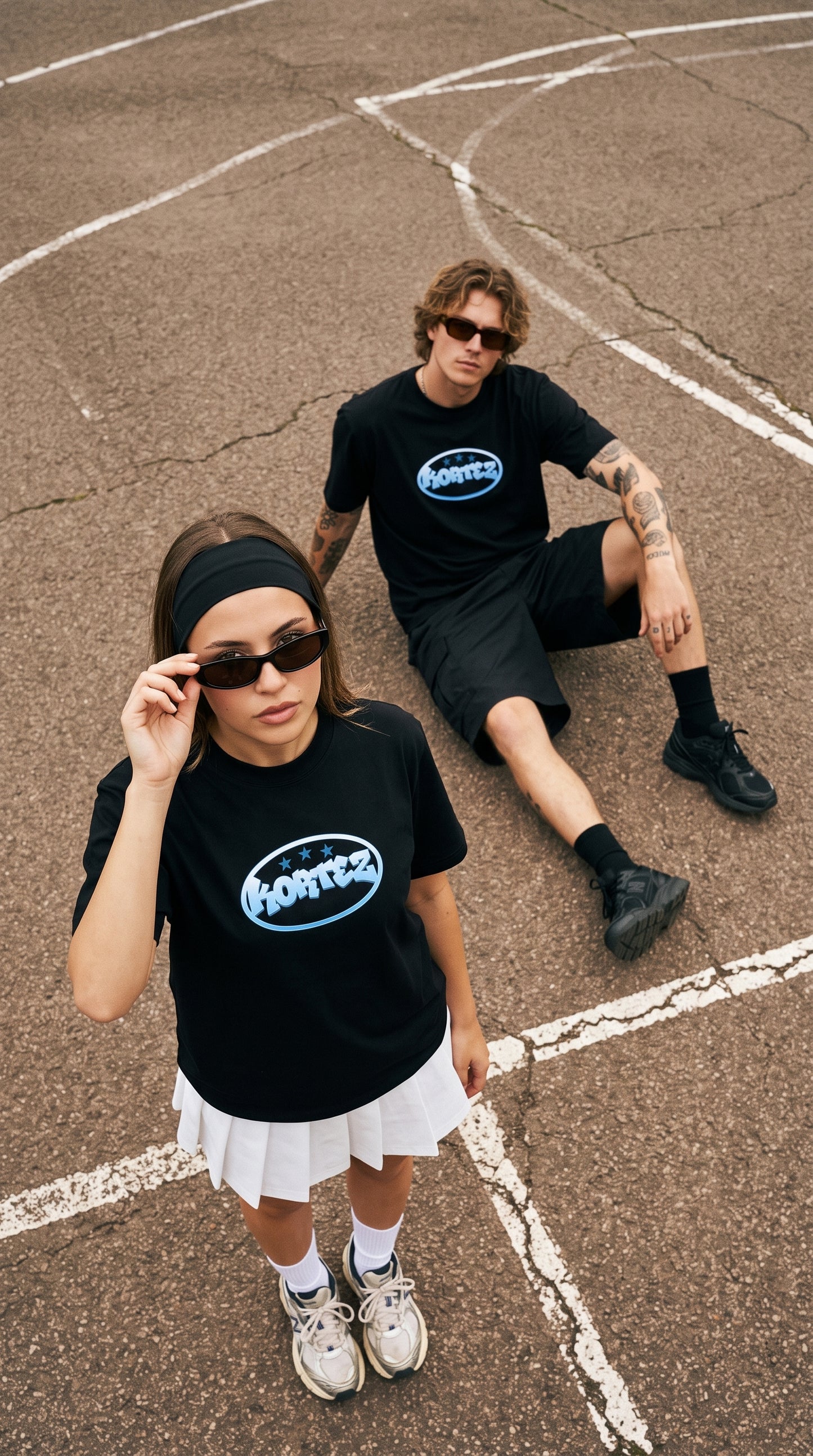 Two models wearing matching black t-shirts with a blue and white 'bombers' logo, styled on an outdoor basketball court. The model in the foreground wears a white pleated skirt and white sneakers, while the model in the background wears black shorts and black sneakers.