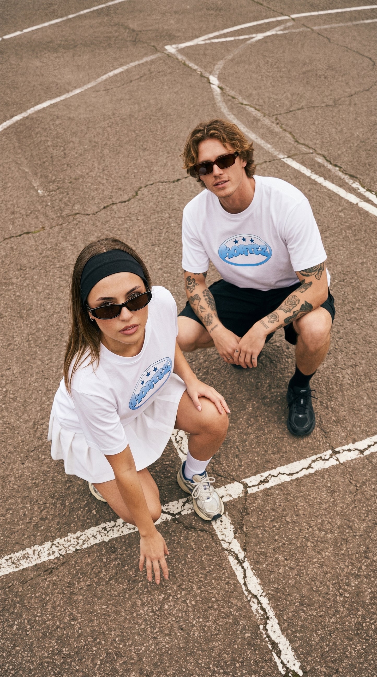 A man and a woman crouching on an outdoor basketball court wearing matching white t-shirts with a blue and white graphic logo on the chest.