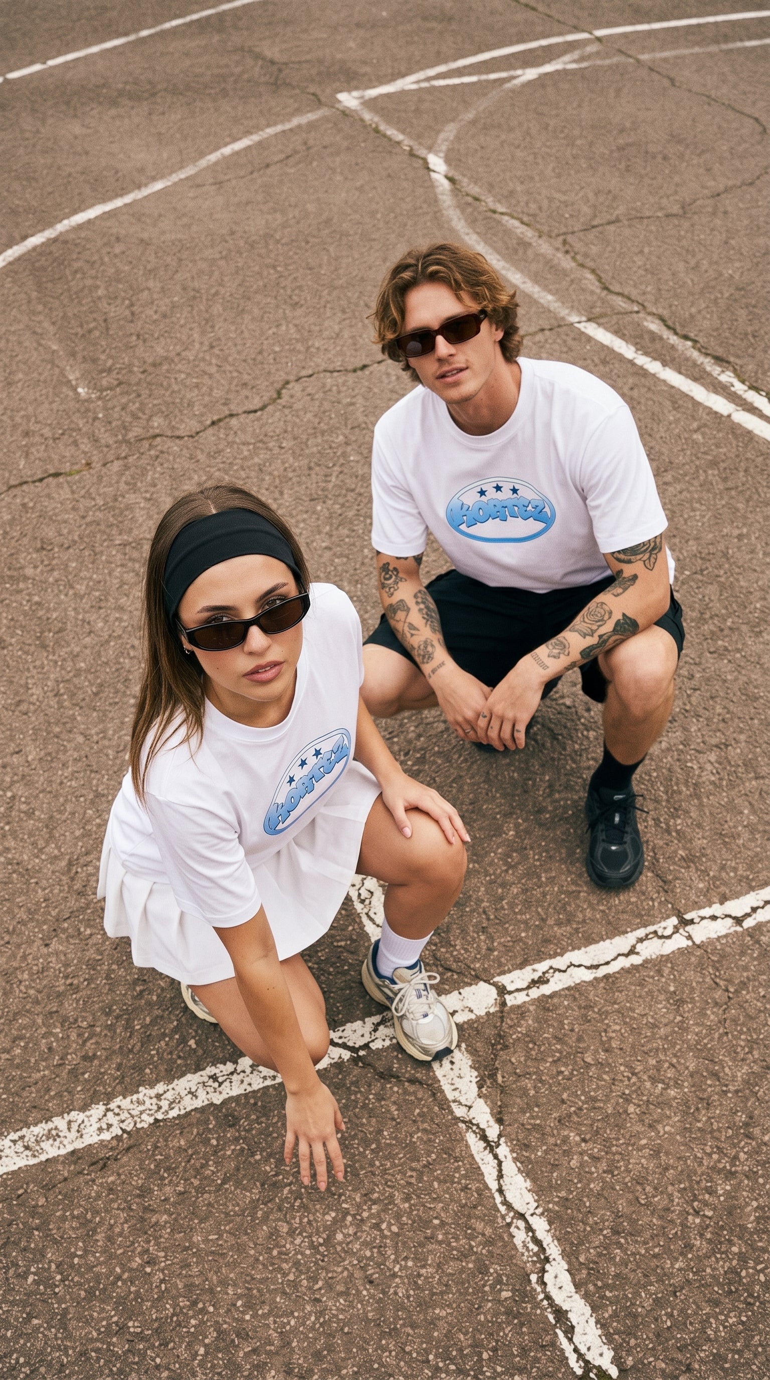 A man and a woman crouching on an outdoor basketball court wearing matching white t-shirts with a blue and white graphic logo on the chest.